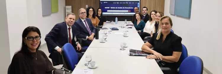 Sala de reunião com mesa longa e pessoas sentadas ao redor. Homens e mulheres, em trajes formais, participam de uma conversa. Alguns anotam, outros escutam com atenção. Ao fundo, uma tela exibe o título “Semana de Saneamento, Precedentes e Ações Coletivas”. O ambiente é claro, com paredes neutras e painéis coloridos.