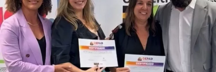 Quatro pessoas estão lado a lado, em pé, sorrindo para a foto. Ao fundo, há um painel colorido com o logotipo do Governo do Estado da Bahia repetido. Duas mulheres no centro seguram certificados com as duas mãos. À esquerda, uma mulher de blazer lilás apoia o certificado de uma delas. À direita, um homem de barba, óculos e blazer cinza coloca o braço em volta do grupo. Todos usam roupas formais. A cena transmite um momento de entrega de certificados.