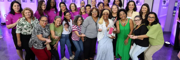 Grupo de mulheres posa para foto em um espaço interno com iluminação roxa. Elas estão lado a lado e também agachadas na frente, sorrindo e olhando para a câmera. No centro, duas mulheres se destacam: uma de blusa cinza com bolinhas e outra com vestido claro e lenço na cabeça, segurando um pequeno presente. As roupas são variadas, com cores como rosa, verde, preto e estampas. Ao fundo, há colunas, quadros na parede e luzes decorativas. O clima é alegre e de celebração.