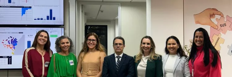 Grupo de sete pessoas posa para foto em um ambiente institucional. Ao fundo, há telas exibindo painéis com gráficos e dados. Os participantes estão alinhados lado a lado, vestidos com roupas formais e crachás de identificação, em frente a um espaço de monitoramento. O ambiente é bem iluminado e organizado, sugerindo uma apresentação ou visita técnica.