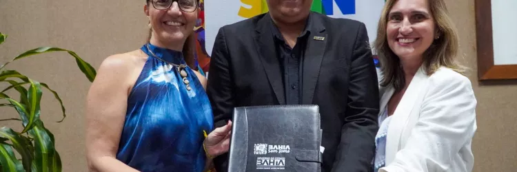 Três pessoas posam sorrindo para a foto em um ambiente interno. Ao centro, um homem de terno preto segura uma pasta preta com as mãos. A pasta tem o texto “Bahia Sem Fome” e “Governo do Estado Bahia”. À esquerda, uma mulher de óculos usa blusa azul sem mangas e segura a pasta junto com ele. À direita, outra mulher, com blazer branco, também segura a pasta. Ao fundo, há um painel colorido com ilustrações de alimentos e uma planta à esquerda.