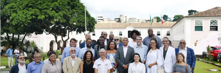 Grupo de cerca de vinte pessoas posa para uma foto ao ar livre. Elas estão organizadas em duas fileiras, em pé, sobre um pequeno palco. A maioria veste roupas sociais, como ternos, camisas e vestidos. Ao fundo há um prédio histórico de dois andares, com paredes claras, janelas verdes e telhado de telhas claras. Bandeiras estão hasteadas no centro do edifício. À esquerda há uma grande árvore com copa cheia e, ao redor, um jardim com gramado. Algumas pessoas aparecem ao fundo caminhando pelo espaço.