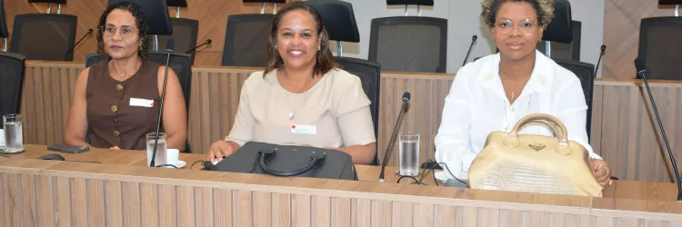 Três mulheres estão sentadas lado a lado em uma mesa de reunião, olhando para a câmera. Sobre a mesa há bolsas, copos de água e equipamentos de áudio. Ao fundo aparecem cadeiras e um painel de madeira na parede.