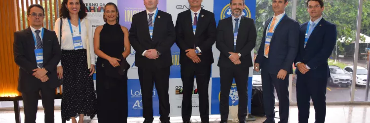 Grupo de pessoas posando para foto em frente ao painel do evento “Caravanas de Inovação”. Todos estão alinhados, vestidos formalmente e com crachás. O ambiente é iluminado, com janelas ao fundo mostrando a área externa.