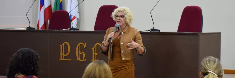 Mulher de cabelo curto e cacheado, loiro claro, está em pé à frente de um auditório, segurando um microfone. Usa óculos e roupa em tom marrom. Ela gesticula com uma das mãos enquanto fala, demonstrando expressão séria e envolvida. Ao fundo, há uma mesa com cadeiras e três bandeiras. Na frente dela, pessoas estão sentadas em cadeiras vermelhas, vistas de costas, assistindo à apresentação. O ambiente é organizado e lembra uma palestra ou evento institucional.