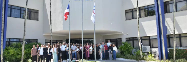Foto externa, em dia ensolarado. Ao centro, um prédio branco com a sigla “PGE” e o nome “Procuradoria Geral do Estado” na fachada. Em frente, três mastros com bandeiras, incluindo a do Brasil. No gramado, as letras “PGE” formadas com pedras. Um grande grupo de pessoas está reunido na entrada, posando para a foto. Há palmeiras altas ao redor e janelas nas laterais do prédio. A cena transmite um encontro institucional.