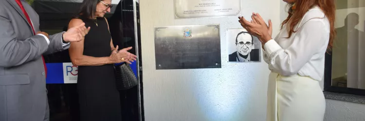 Três pessoas em um evento oficial. À esquerda, um homem de terno cinza gesticula; ao centro, uma mulher de vestido preto aplaude; à direita, uma mulher com camisa branca e lenço também aplaude. Eles estão diante de placas de inauguração na parede.