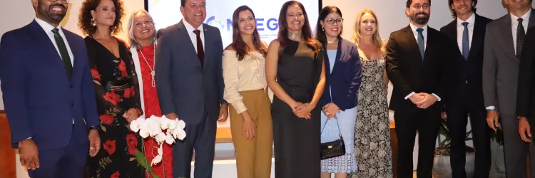 Grupo de várias pessoas alinhadas para uma foto em evento formal. Homens e mulheres vestem roupas sociais e estão sorrindo. Ao fundo, há um telão com logotipo e decoração simples, indicando uma cerimônia ou encontro institucional.