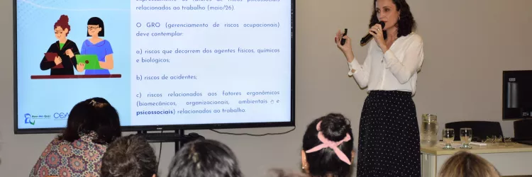 Uma mulher apresenta conteúdo exibido na tela, segurando um microfone. O slide mostra informações sobre normas e riscos no trabalho. Parte do público aparece de costas, acompanhando atentamente a explicação.