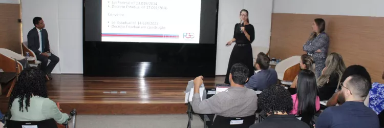 Uma palestrante de vestido preto fala ao microfone no palco, apontando para um telão que detalha informações sobre "LEGISLAÇÃO" e "PRESTAÇÃO DE CONTAS". Dois outros palestrantes acompanham a fala nas laterais do palco.