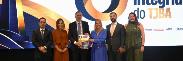 Seis pessoas estão em pé, lado a lado, diante de um grande painel iluminado. Ao fundo, aparece a imagem de um farol, um símbolo circular em azul e laranja e o texto “Encontro de Integridade do TJBA”. As pessoas usam roupas sociais; duas vestem ternos, três usam vestidos e uma usa blusa e calça. A pessoa ao centro segura um certificado ou material do evento. Todos posam para a foto.