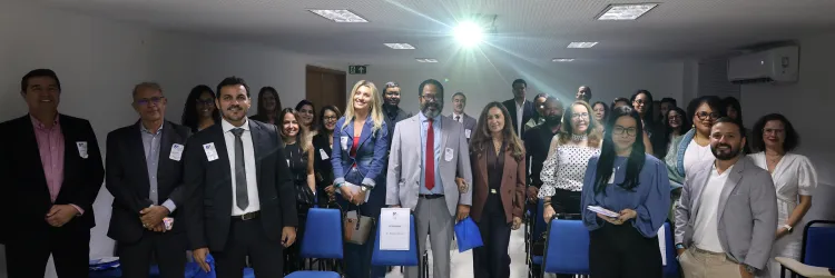 Fotografia de um grupo numeroso de pessoas reunidas em uma sala. Elas estão em pé e voltadas para a câmera, muitas sorrindo. A maioria usa roupas sociais, como ternos, blazers, camisas e vestidos, e algumas usam crachás. Ao centro, há um homem de terno cinza e gravata vermelha. À frente, há cadeiras azuis. No canto inferior esquerdo, uma mesa coberta com toalha branca exibe garrafas de água e taças. O ambiente tem teto com iluminação embutida e ar-condicionado.