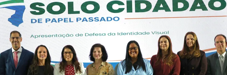 Em frente a um painel com o texto “Defensoria Pública – BA” e “Solo Cidadão – De Papel Passado”, sete pessoas posam para uma foto. Da esquerda para a direita, há três homens e quatro mulheres, vestidos formalmente. As mulheres usam roupas em preto, branco, vermelho, bege e azul-claro. Todos estão de pé, lado a lado, sorrindo ou olhando para a câmera. O painel também traz a frase “Apresentação de Defesa da Identidade Visual”.