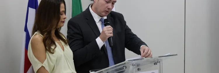 Um homem de terno preto segura um microfone e lê um documento diante de um púlpito de acrílico com o logotipo da PGE-BA. Ao lado, uma mulher de blusa clara e calça verde acompanha o momento. Ao fundo, estão bandeiras institucionais. A cena ocorre em uma sala de ambiente formal.