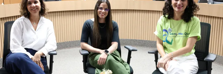 Fotografia de três mulheres sentadas em cadeiras, em um ambiente institucional. A mulher à esquerda usa camisa branca, calça azul-marinho e sapatos marrons. A mulher ao centro veste blusa preta, calça verde, óculos e relógio. A mulher à direita usa camiseta verde-clara com estampa, calça branca e tênis. Todas sorriem para a câmera. À frente delas, há uma mesa de vidro com microfones e um pequeno arranjo de flores. Ao fundo, vê-se um painel de madeira com a identificação do TCE-BA