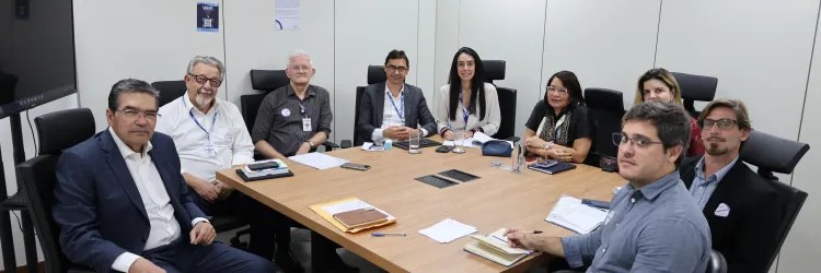 Sala de reunião com paredes claras, mesa retangular de madeira e cadeiras pretas. Nove pessoas estão sentadas ao redor da mesa, voltadas para a câmera. Algumas usam crachás e estão com papéis, cadernos, celulares e canetas sobre a mesa. Ao fundo, há uma tela de televisão, cartazes nas paredes e luminárias no teto.