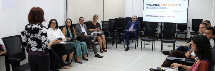 Fotografia de uma reunião em uma sala ampla e clara, com pessoas sentadas em cadeiras dispostas em semicírculo. Ao centro, uma mulher está em pé, de costas para a câmera, conduzindo a atividade. Os participantes fazem anotações e utilizam computadores. Ao fundo, há uma tela exibindo a apresentação “Valores e Impacto no Ecossistema” e informações sobre mentoria executiva em liderança.