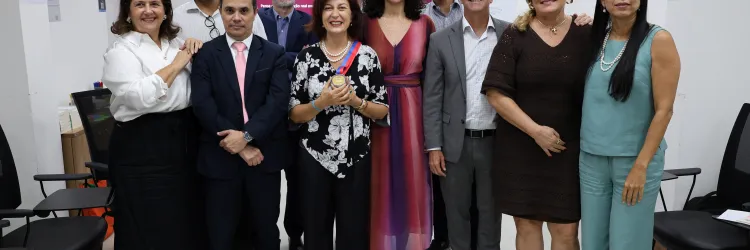 Fotografia de um grupo de onze pessoas reunidas em uma sala. Elas estão em pé, lado a lado, sorrindo e olhando para a câmera. Ao centro, uma mulher usa blusa preta com estampa de flores brancas, colar de pérolas e segura uma medalha. Os demais participantes usam roupas sociais e casuais. Ao fundo, há uma tela com uma apresentação e cadeiras organizadas pela sala.