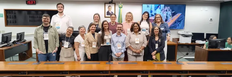 Foto de um grupo de cerca de 14 pessoas reunidas em uma sala de reuniões. Elas estão de pé, lado a lado, atrás de uma longa mesa de madeira com microfones. Ao fundo, há bandeiras, um retrato, um relógio digital e uma tela exibindo outra imagem da sala. Todos posam para a foto e usam crachás de identificação.