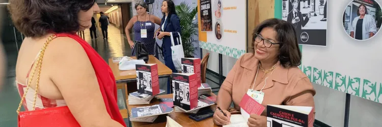Foto de um estande de evento. À direita, uma mulher de óculos, usando roupa social em tom salmão e crachá, está sentada à mesa, sorrindo e autografando um livro. À esquerda, outra mulher, de costas para a câmera, usa roupa vermelha e carrega uma bolsa da mesma cor. Sobre a mesa, há vários exemplares do livro “Casos e causos da PGE-BA: entre prazos, cafezinhos e risadas”. Ao fundo, outras pessoas conversam e há painéis com materiais de divulgação.