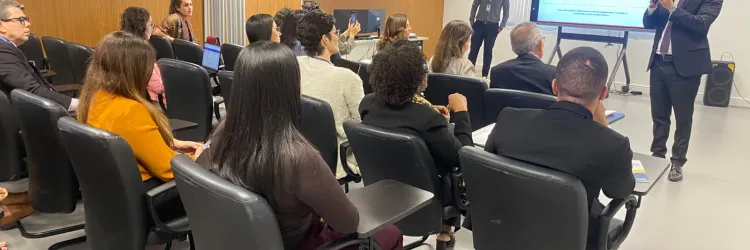 Sala de reunião com várias pessoas sentadas em cadeiras diante de uma tela. Ao fundo, um homem de terno segura um microfone e fala para o grupo. Outro homem está em pé próximo à tela, que exibe uma apresentação. As paredes têm revestimento de madeira, há janelas com persianas, ar-condicionado e projetor no teto. Os participantes observam a apresentação e alguns usam celulares ou computadores.