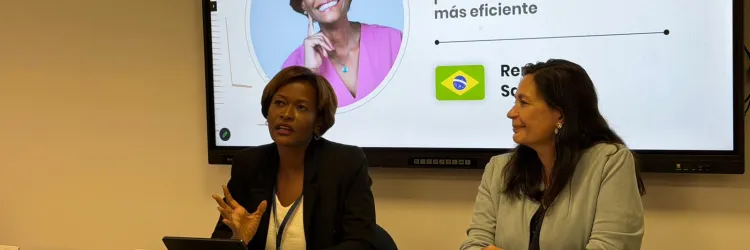 Fotografia de um painel de evento. Duas mulheres estão sentadas à mesa. À esquerda, uma mulher negra, de cabelos curtos, usa blazer preto e blusa clara. À direita, uma mulher de cabelos longos usa blusa cinza. Ao fundo, um telão exibe o título “Boa administração na era digital: Governança de dados para uma gestão pública mais eficiente”, além das imagens e nomes das participantes. Sobre a mesa, há placas de identificação, microfones e objetos pessoais.