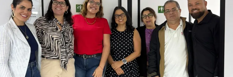 Foto de sete pessoas em pé, lado a lado, em um ambiente com divisórias brancas e estruturas pretas. Da esquerda para a direita, são cinco mulheres e dois homens. As pessoas usam roupas casuais e sociais, em diferentes cores e estampas, e olham para a câmera, sorrindo. Ao fundo, há portas ou divisórias com placas verdes de sinalização. No canto inferior esquerdo, aparecem cadeiras azuis. O piso é claro e o teto tem placas brancas com estrutura preta.