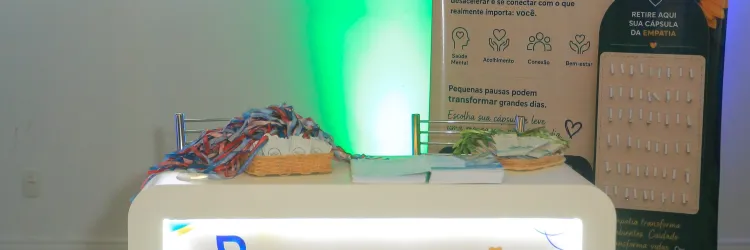 Foto de um espaço de divulgação chamado “Estação Empatia”. Ao centro, há um balcão iluminado, com cartazes sobre empatia, acolhimento, conexão e bem-estar. Sobre o balcão, estão cestas com materiais e pequenos cartões. Ao fundo, um painel vertical traz o mesmo tema, com uma flor, folhas e um suporte com cápsulas de papel. O ambiente é interno, com paredes claras e iluminação decorativa em verde e azul.