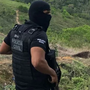 Agente da Polícia Civil usando colete tático e balaclava, segurando uma arma, em uma área rural com vegetação e morros ao fundo, durante uma operação
