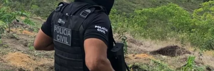 Agente da Polícia Civil usando colete tático e balaclava, segurando uma arma, em uma área rural com vegetação e morros ao fundo, durante uma operação