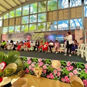A imagem mostra um evento realizado em um espaço coberto com um palco elevado. Sobre o palco, há várias pessoas sentadas em cadeiras de plástico brancas, e uma pessoa de pé fala ao microfone. O banner ao fundo indica que o evento é relacionado ao "Encontro Nacional de Mulheres do MPA" 