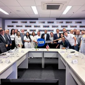 Um grupo de pessoas reunido em uma sala de reunião, posando para uma foto em frente a uma mesa branca com papéis e copos d'água. Ao fundo, há um banner com o logotipo do Banco do Brasil e bandeiras do Brasil e da Bahia