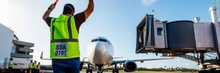  A imagem mostra um funcionário do aeroporto, usando um colete de alta visibilidade verde com detalhes refletivos e segurando dois bastões iluminados (provavelmente usados para sinalização), orientando um avião em direção ao portão de embarque ou desembarque. O avião está na pista, posicionado em frente a uma ponte de embarque, conhecida como finger