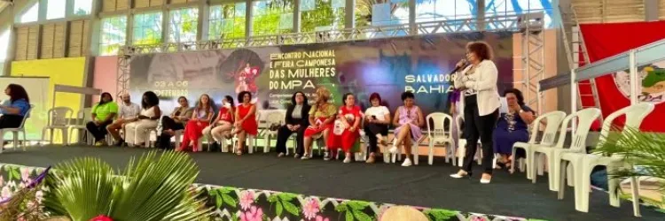 A imagem mostra um evento realizado em um espaço coberto com um palco elevado. Sobre o palco, há várias pessoas sentadas em cadeiras de plástico brancas, e uma pessoa de pé fala ao microfone. O banner ao fundo indica que o evento é relacionado ao "Encontro Nacional de Mulheres do MPA" 