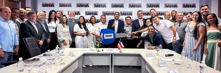 Um grupo de pessoas reunido em uma sala de reunião, posando para uma foto em frente a uma mesa branca com papéis e copos d'água. Ao fundo, há um banner com o logotipo do Banco do Brasil e bandeiras do Brasil e da Bahia