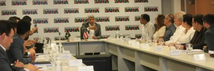 A imagem mostra uma reunião em uma sala de conferências com várias pessoas sentadas em torno de uma mesa em formato de "U"