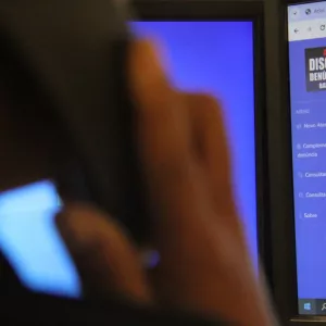 Uma pessoa simula uma ligação e ao fundo está o site do Disque Denúncia