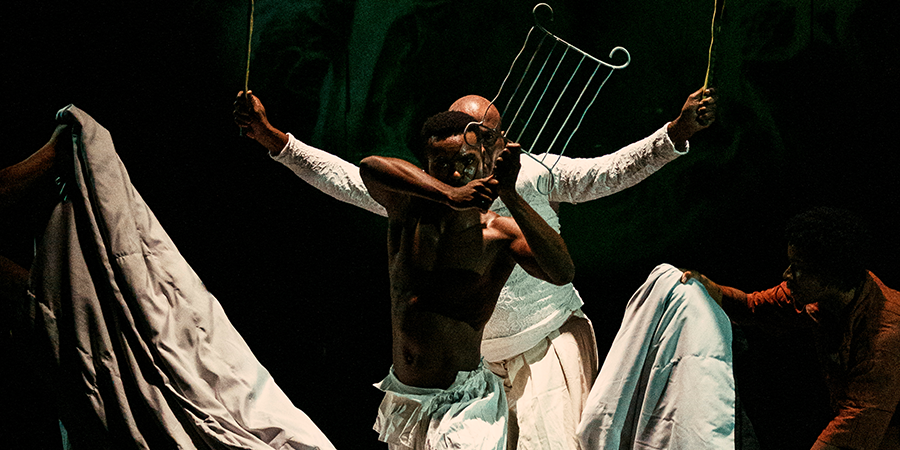 Imagem enquadra um rapaz negro, à frente, que segura um indumentária do orixá Iroko chamado "Orelha do Tempo" (ou grelha). Atrás dele, um homem negro mais velho está enquadrado apenas com a metade da cabeça, com o olhar por trás do rapaz e ele segura, nas duas mãos, folhas da espada de Ogum.