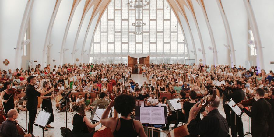 Imagem enquadra músicos da OSBA posicionados em um palco para frente de um público de pessoas sentadas em bancos dentro de um Santuário 