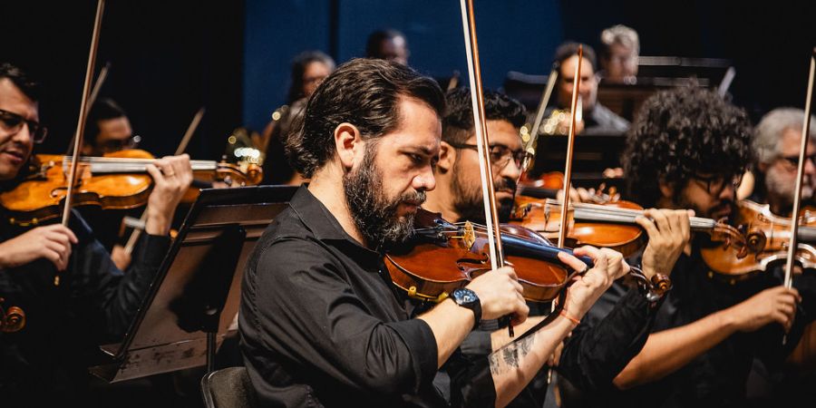 Imagem centraliza, ao meio, um músico com um violino. Ele é um homem branco, de barba, e um relógio no braço. Ele está de perfil. Em volta dele estão outros músicos, homens, que também estão com violinos. Todos usam camisa social preta.