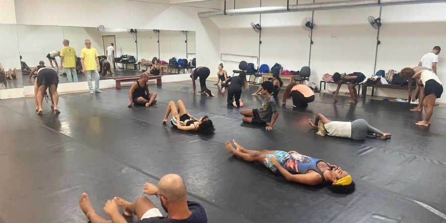 Imagem mostra um grupo de pessoas reunidas em uma sala de ensaio de dança. Algumas estão deitadas, outras estão em posição de alongamento.