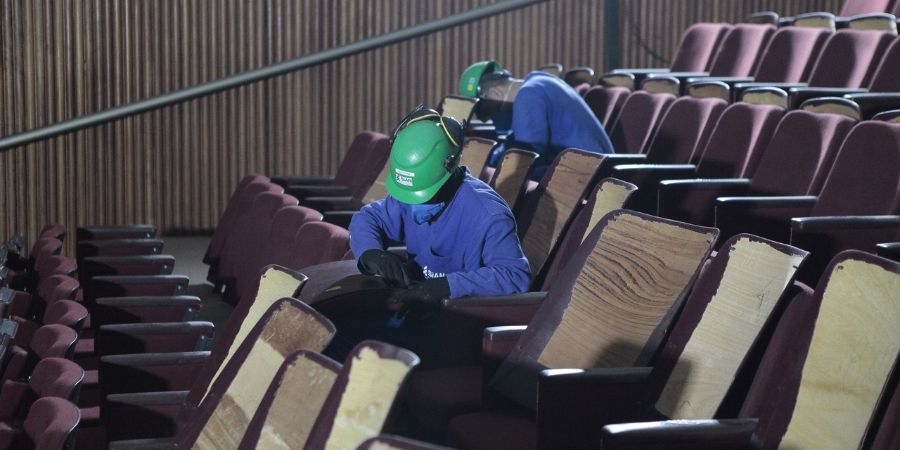 Imagem enquadra dois trabalhadores que utilizam capacetes verdes. Eles estão retirando poltronas da Sala Principal do Teatro Castro Alves