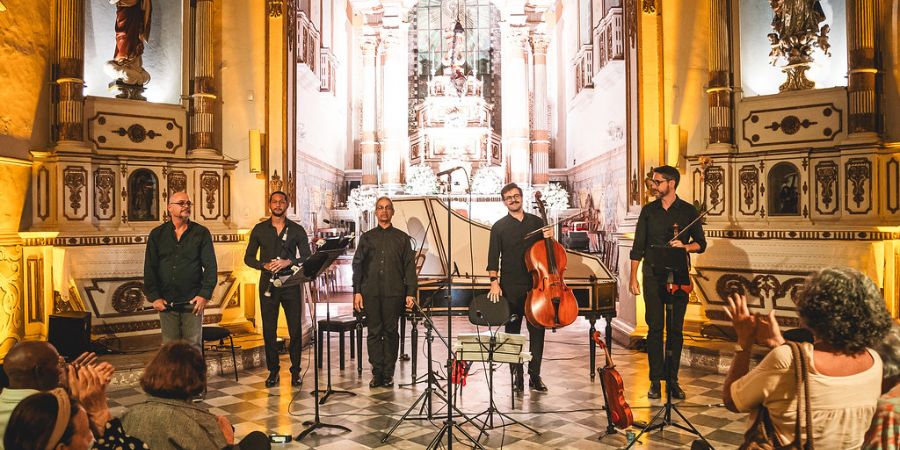 Esta é uma fotografia que retrata um quinteto se apresentando com instrumentos da música clássica, como violino, violoncelo e flautas, dentro de uma igreja ou capela de estilo barroco. O crédito da foto é de Caio Lírio.