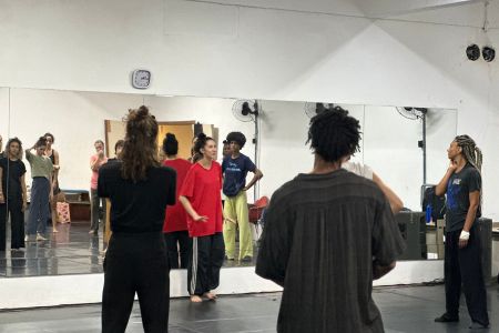 Em uma sala de ensaio de dança com espelhos ao longo da parede, um grupo de pessoas em roupas confortáveis acompanha uma instrução. Em primeiro plano, duas pessoas aparecem de costas, observando os demais. Ao fundo, uma mulher de camiseta vermelha larga e calça preta fala ao centro, enquanto outros bailarinos ao redor e refletidos no espelho prestam atenção.