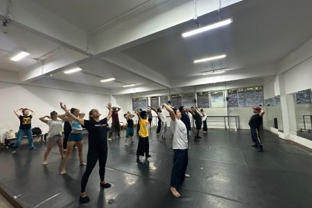Em uma ampla sala de dança com piso preto e paredes brancas, vários dançarinos realizam um alongamento coletivo. Espalhados pelo espaço sob a luz de luminárias no teto, eles estão em pé, de pernas afastadas, inclinando o tronco e erguendo os braços. Ao fundo, à direita, um homem de boné observa o grupo.