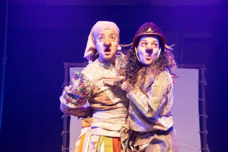 Duas atrizes em uma peça de teatro no palco com iluminação roxa, vistas do meio do corpo para cima. Elas usam figurinos retalhados e narizes postiços, expressando espanto. À esquerda, uma delas usa um lenço claro na cabeça e tem a boca aberta em surpresa. À direita, a outra, de cabelos cacheados e chapéu escuro, segura o braço da companheira enquanto olha para a frente com olhos arregalados. Ao fundo, vê-se um cenário de tecido branco.