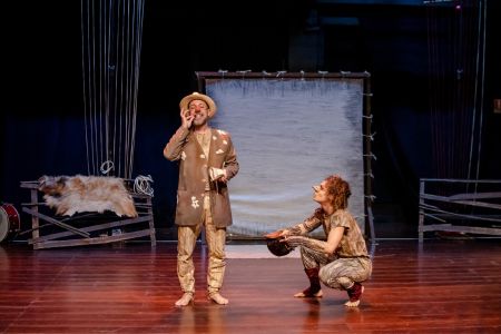 No palco de um teatro, sob iluminação suave, dois atores encenam uma peça. À esquerda, um homem de chapéu de palha, nariz de palhaço vermelho e terno marrom remendado está em pé, sorrindo e levando a mão ao rosto. À direita, outra pessoa, com nariz postiço pontudo e roupa em tons de terra está agachada, olhando para ele de perfil e segurando um chapéu escuro. Ao fundo, um painel de tecido claro e, nas laterais, estruturas de madeira com cordas e peles cenográficas.