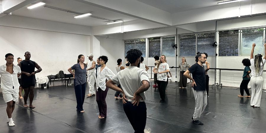 Grupo de bailarinos em uma ampla sala de ensaio com piso preto linóleo e paredes brancas iluminadas por luzes de led. Os dançarinos vestem roupas confortáveis e casuais em tons neutros — como branco, preto e cinza — e estão espalhados pelo espaço, alguns de costas e outros de frente, praticando movimentos corporais. Ao fundo, observam-se grandes janelas e barras de dança encostadas na parede.