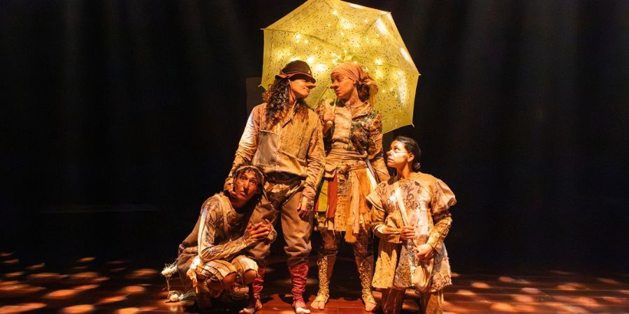 Quatro atores em uma peça de teatro, vestidos com roupas retalhadas em tons de terra, no palco escuro. No centro, dois deles, com narizes postiços, estão sob um guarda-chuva amarelo iluminado por pequenas luzes. Uma terceira atriz, agachada à esquerda, e uma atriz em pé à direita olham atentamente para cima.