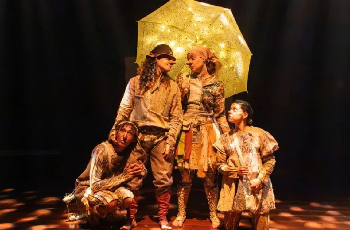 Quatro atores em uma peça de teatro, vestidos com roupas retalhadas em tons de terra, no palco escuro. No centro, dois deles, com narizes postiços, estão sob um guarda-chuva amarelo iluminado por pequenas luzes. Uma terceira atriz, agachada à esquerda, e uma atriz em pé à direita olham atentamente para cima.